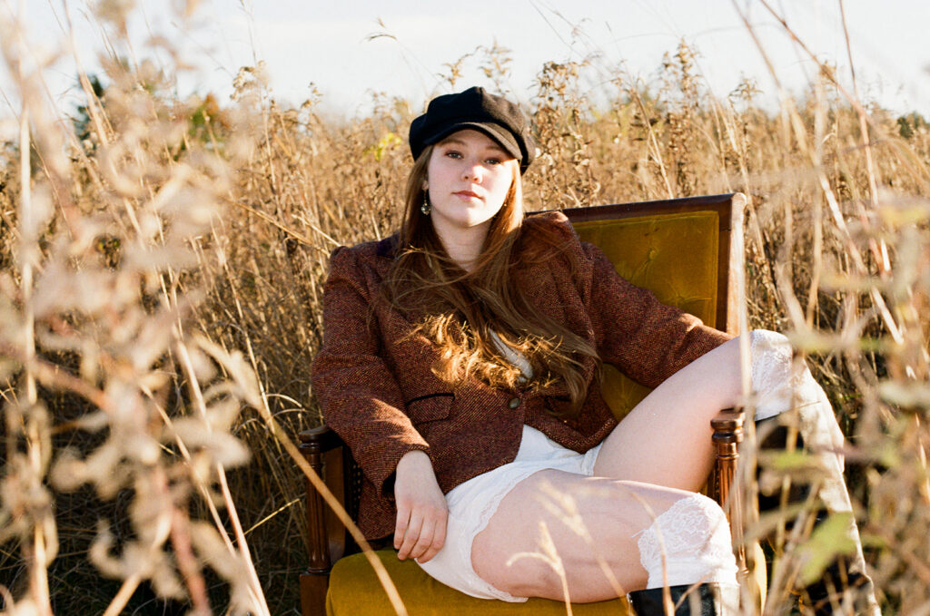 A young woman wearing a newsboy cap is seated on a mustard yellow vintage chair for her Minnesota senior photos 