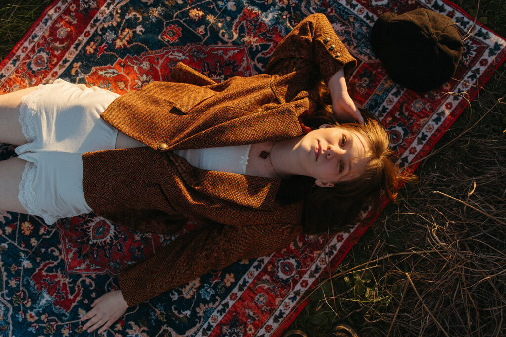 A golden hour Minnesota senior picture depicts a young woman in a brown tweed blazer lying atop a vintage rug at Afton State Park