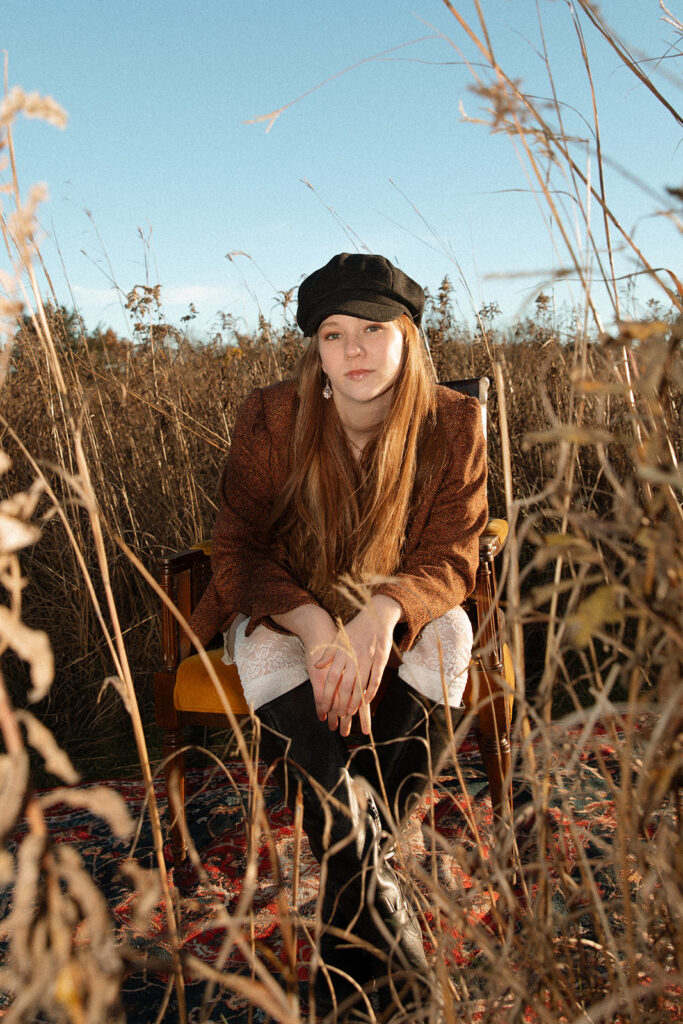 A high school senior leans forward in a vintage chair surrounded by prairie grass during Minnesota senior pictures at Afton State Park 